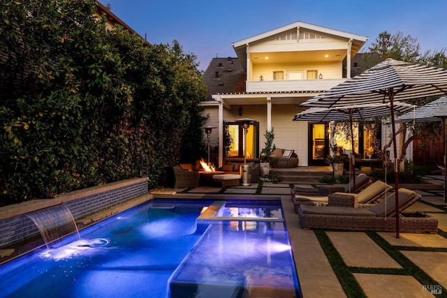 back of property featuring a balcony, a patio area, a pool with connected hot tub, and an outdoor living space with a fire pit