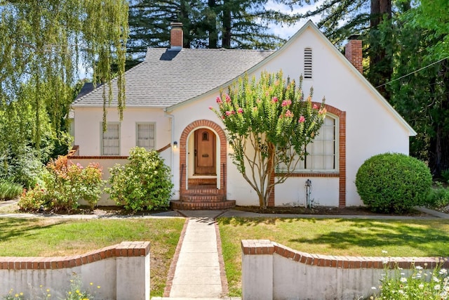 english style home featuring a front yard, roof with shingles, a chimney, and stucco siding