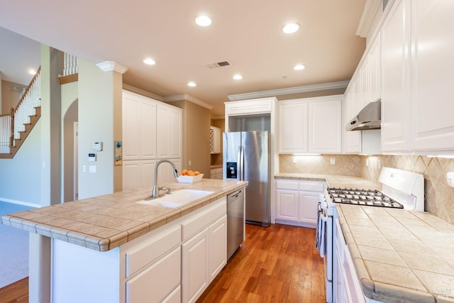 kitchen with tile counters, white cabinets, wall chimney exhaust hood, appliances with stainless steel finishes, and a sink