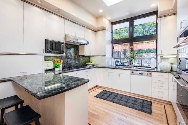 kitchen featuring under cabinet range hood, a peninsula, white cabinets, dishwasher, and dark stone countertops