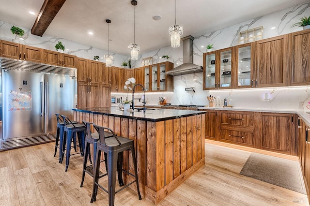 kitchen with stainless steel appliances, a kitchen island with sink, brown cabinets, and wall chimney exhaust hood