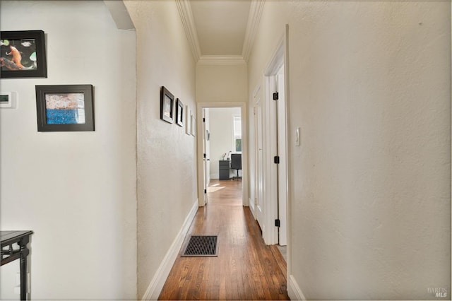 hall featuring a textured wall, wood finished floors, visible vents, baseboards, and ornamental molding