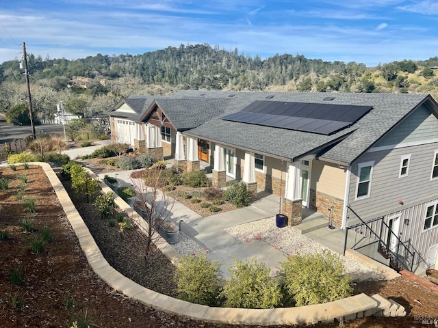 modern farmhouse style home featuring a forest view, stone siding, solar panels, and roof with shingles