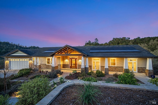view of front facade featuring stone siding, covered porch, an attached garage, a shingled roof, and roof mounted solar panels
