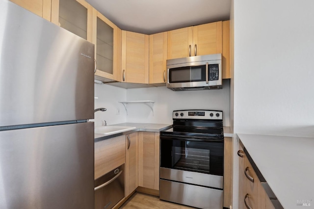 kitchen featuring glass insert cabinets, appliances with stainless steel finishes, light countertops, light brown cabinetry, and a sink