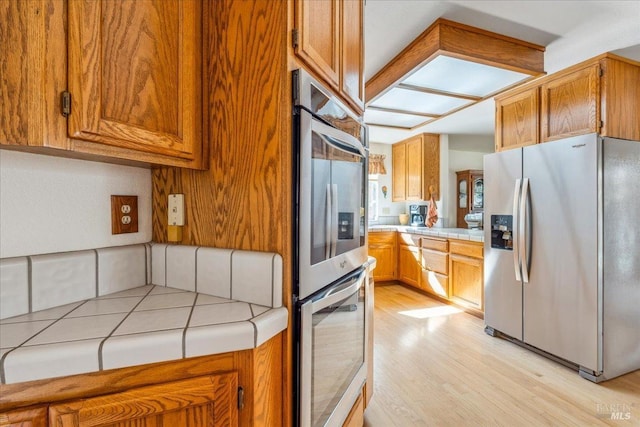 kitchen featuring stainless steel appliances, light wood-style flooring, tile counters, and brown cabinets