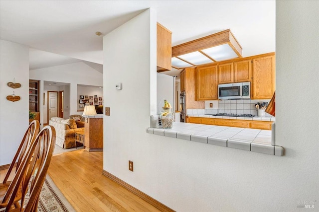 kitchen featuring tile countertops, stainless steel microwave, vaulted ceiling, light wood-style floors, and black gas stovetop