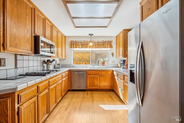 kitchen with brown cabinetry, tile countertops, stainless steel appliances, light wood-type flooring, and backsplash