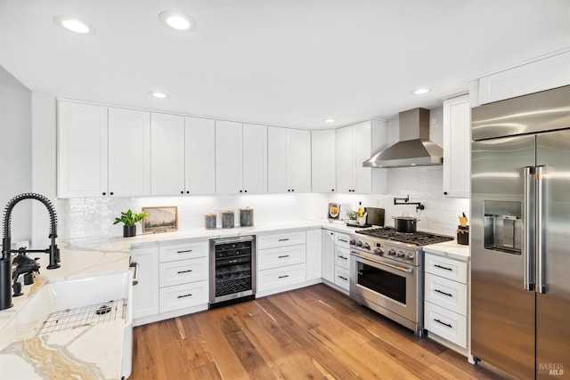 kitchen with light stone counters, a sink, high end appliances, beverage cooler, and wall chimney exhaust hood