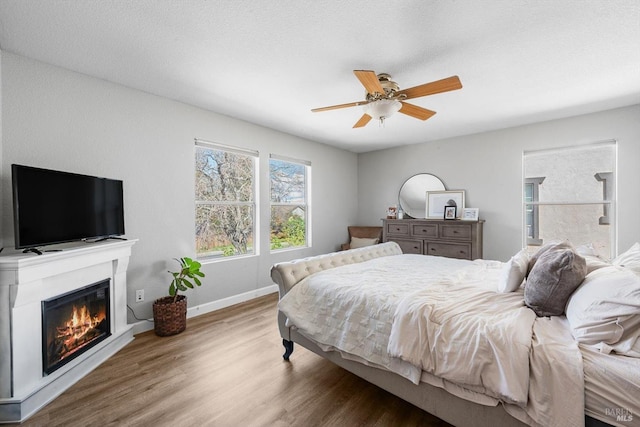 bedroom with a glass covered fireplace, wood finished floors, baseboards, and a ceiling fan