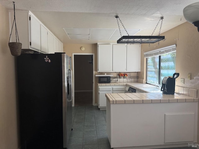 kitchen with tile countertops, black microwave, a peninsula, stainless steel refrigerator with ice dispenser, and white cabinetry