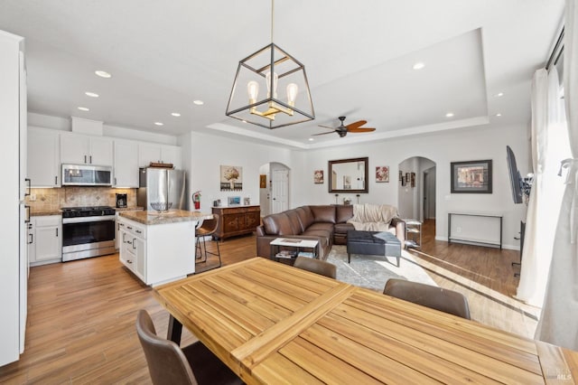 dining room with arched walkways, recessed lighting, a raised ceiling, light wood-style floors, and ceiling fan with notable chandelier