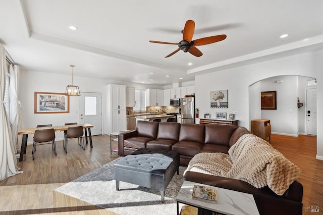 living room with arched walkways, a raised ceiling, ceiling fan with notable chandelier, light wood-style floors, and recessed lighting