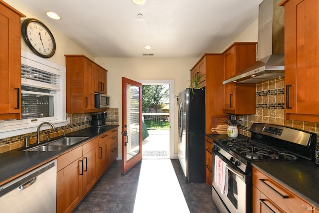 kitchen with brown cabinetry, dark countertops, appliances with stainless steel finishes, wall chimney range hood, and a sink