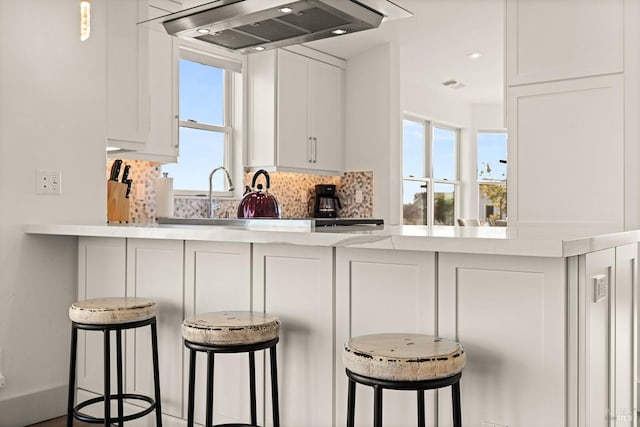 kitchen with a sink, backsplash, white cabinetry, a breakfast bar area, and wall chimney exhaust hood