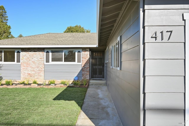 view of exterior entry with a yard, a shingled roof, and brick siding