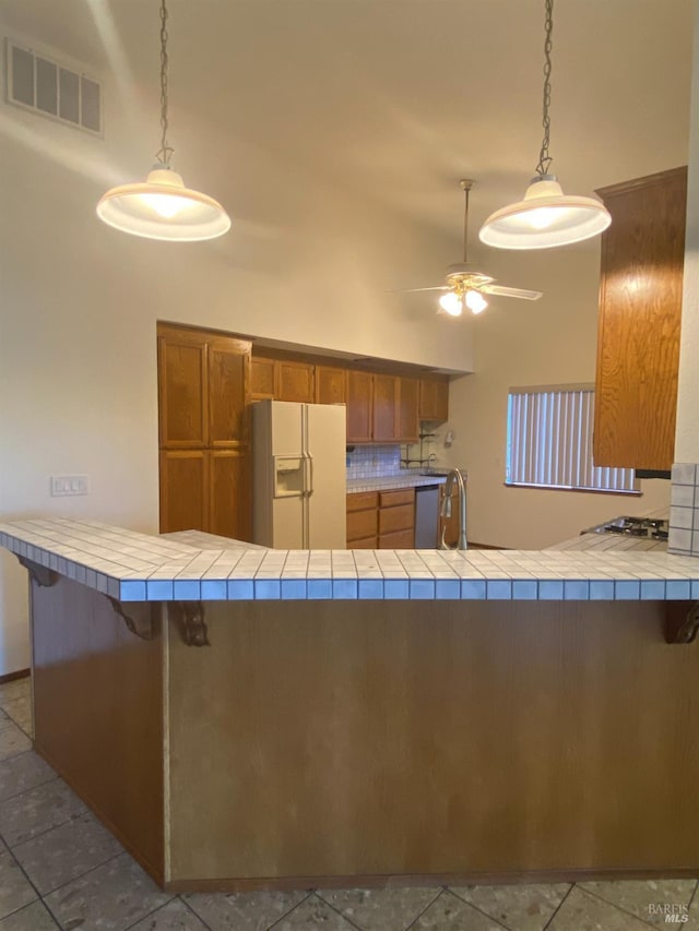 kitchen featuring brown cabinetry, tile counters, white refrigerator with ice dispenser, and visible vents