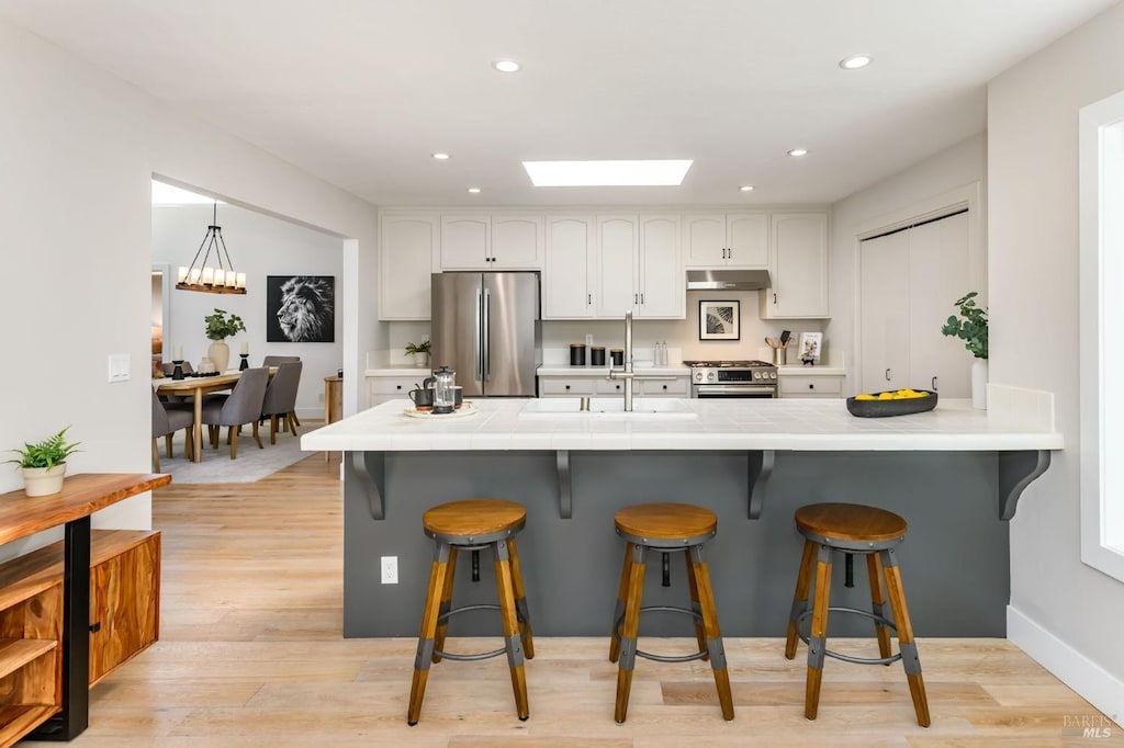 kitchen with a kitchen breakfast bar, under cabinet range hood, white cabinets, a sink, and appliances with stainless steel finishes