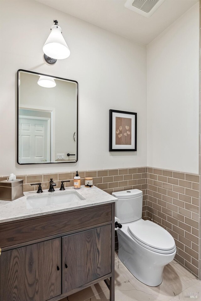 half bathroom featuring vanity, tile walls, toilet, visible vents, and wainscoting