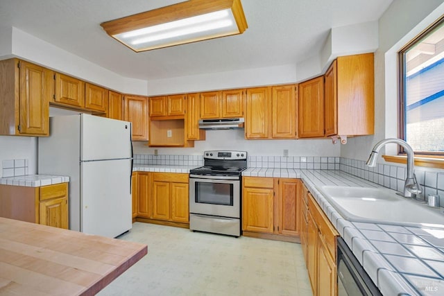 kitchen featuring under cabinet range hood, a sink, stainless steel range with electric cooktop, tile counters, and freestanding refrigerator