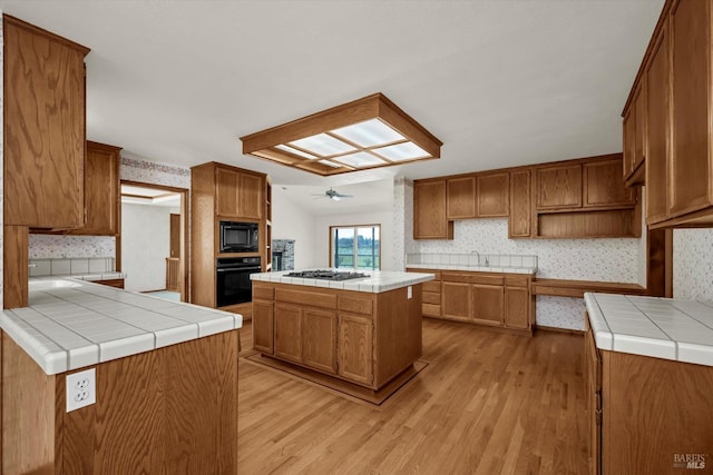 kitchen with a sink, light wood-type flooring, tile counters, black appliances, and wallpapered walls