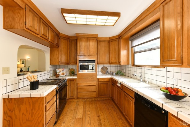 kitchen featuring light wood finished floors, tile counters, brown cabinetry, black appliances, and a sink