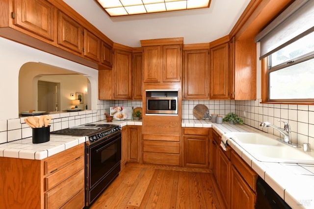 kitchen featuring tile countertops, a sink, light wood-type flooring, black appliances, and backsplash