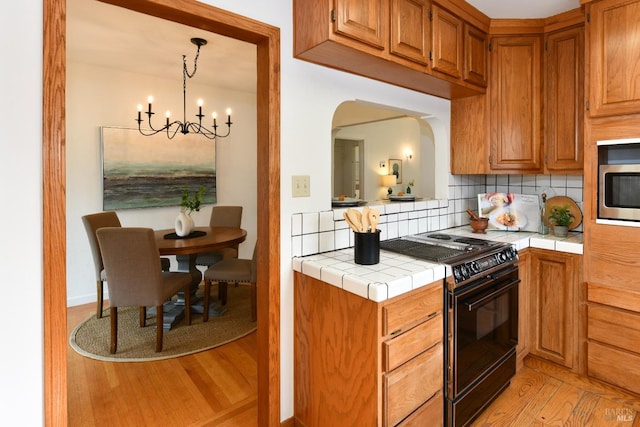 kitchen with black electric range, a notable chandelier, tile countertops, stainless steel microwave, and backsplash