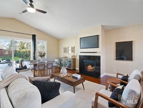 living area with a glass covered fireplace, ceiling fan, vaulted ceiling, wood finished floors, and baseboards