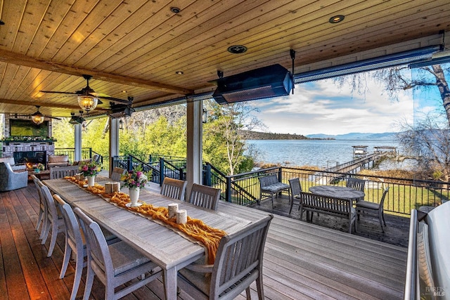 wooden deck featuring a ceiling fan, outdoor dining space, a warm lit fireplace, and a water view