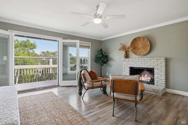 living area with baseboards, a ceiling fan, wood finished floors, crown molding, and a brick fireplace