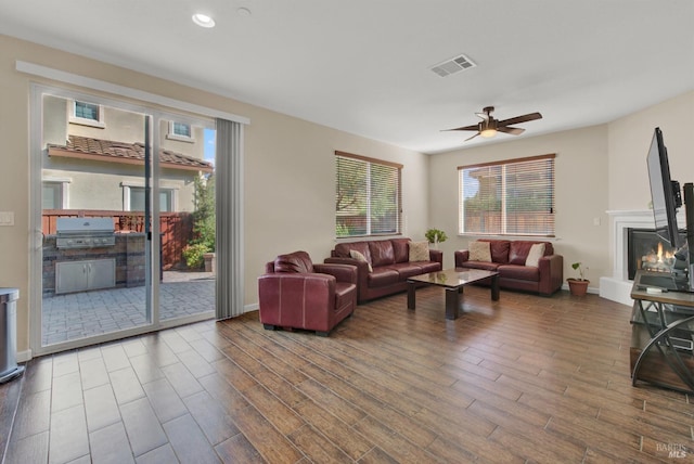 living room with visible vents, a ceiling fan, a glass covered fireplace, wood finished floors, and baseboards