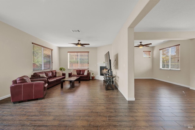 living area featuring a healthy amount of sunlight, a warm lit fireplace, ceiling fan, and dark wood-style flooring