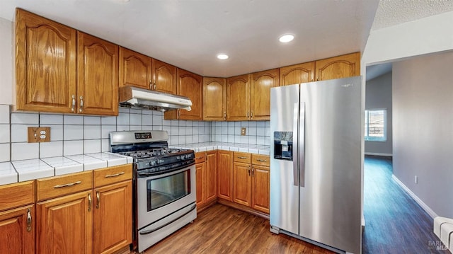 kitchen featuring brown cabinets, under cabinet range hood, tile counters, and stainless steel appliances