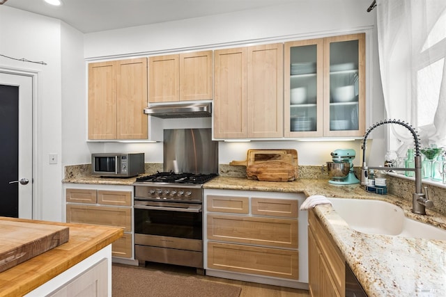 kitchen with appliances with stainless steel finishes, glass insert cabinets, light brown cabinets, a sink, and under cabinet range hood