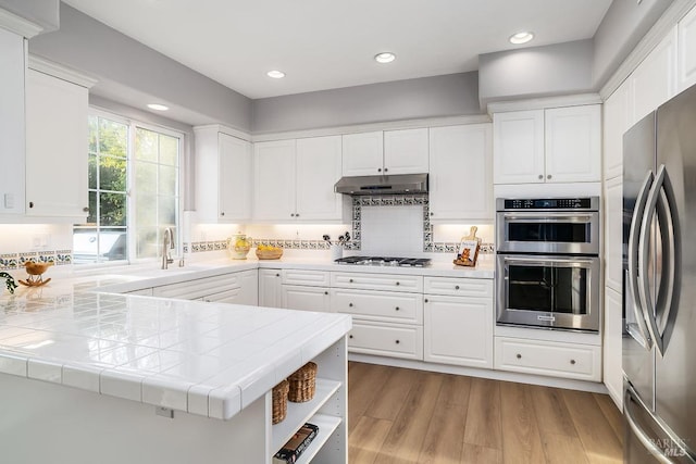 kitchen with under cabinet range hood, stainless steel appliances, a sink, tile counters, and light wood finished floors