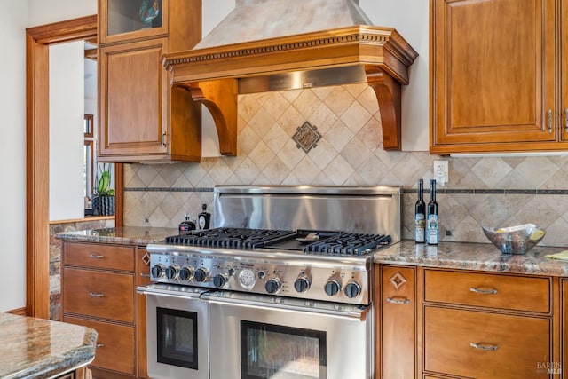 kitchen with stone counters, premium range hood, double oven range, decorative backsplash, and brown cabinetry