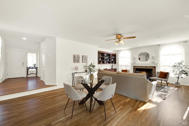 dining space featuring a warm lit fireplace, ceiling fan, wood finished floors, and recessed lighting