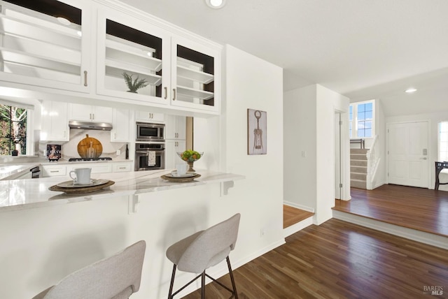 kitchen featuring a breakfast bar, appliances with stainless steel finishes, glass insert cabinets, white cabinetry, and under cabinet range hood