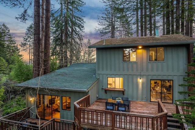 back of property at dusk featuring a deck, a shingled roof, board and batten siding, and outdoor dining area