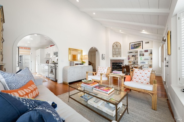 living room with arched walkways, beam ceiling, a brick fireplace, high vaulted ceiling, and light wood-type flooring