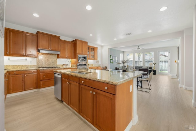 kitchen featuring light stone counters, under cabinet range hood, stainless steel appliances, light wood-type flooring, and brown cabinetry