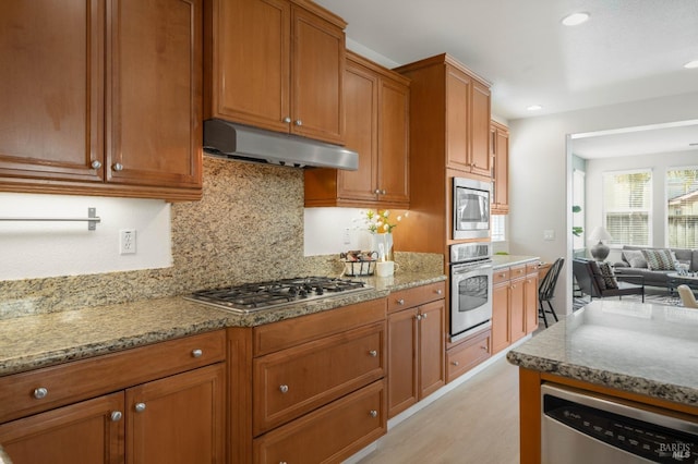 kitchen featuring appliances with stainless steel finishes, brown cabinets, light stone counters, and under cabinet range hood