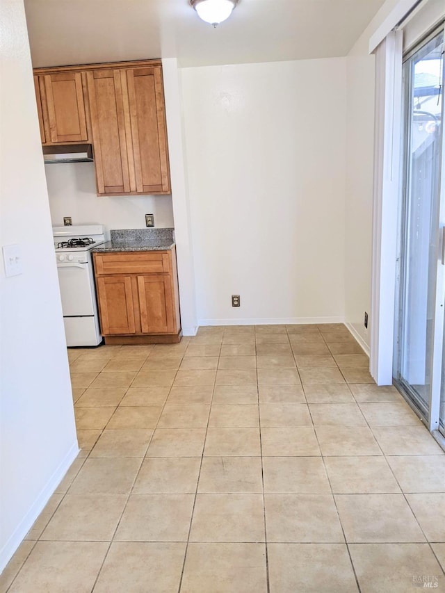 kitchen featuring light tile patterned floors, baseboards, brown cabinetry, under cabinet range hood, and gas range gas stove