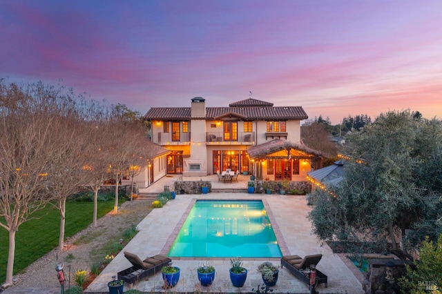 back of house at dusk with a patio, a balcony, stucco siding, a chimney, and a lawn