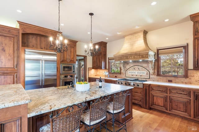 kitchen with light stone counters, premium range hood, light wood finished floors, built in appliances, and backsplash