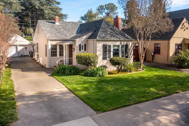 tudor home with roof with shingles, stucco siding, a front yard, an outbuilding, and a chimney