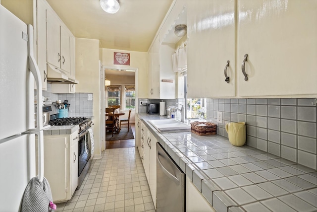kitchen with backsplash, a sink, under cabinet range hood, tile counters, and stainless steel appliances
