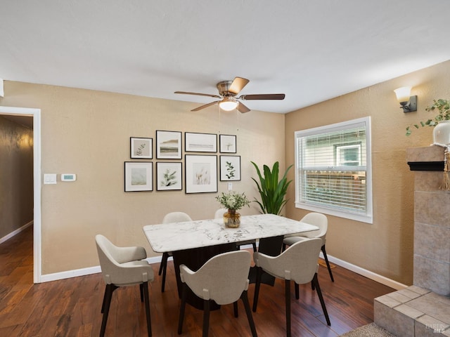 dining room with ceiling fan, a tiled fireplace, baseboards, and wood finished floors