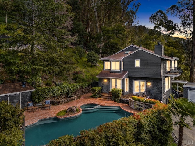 back of property at dusk with a balcony, a patio area, a pool with connected hot tub, and a chimney